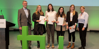 Guido Baranowski (Vorstandsvorsitzender GdF, l.) und Prof. Wiebke Möhring (Prorektorin Studium, r.) stehen auf der Absolventenfeier neben Kathrin Stäblein, Lea Sennekamp, Imren Icel und Lisa Ruffer (v. l.). Im Vordergrund ein großes plastisches Logo der TU Dortmund.