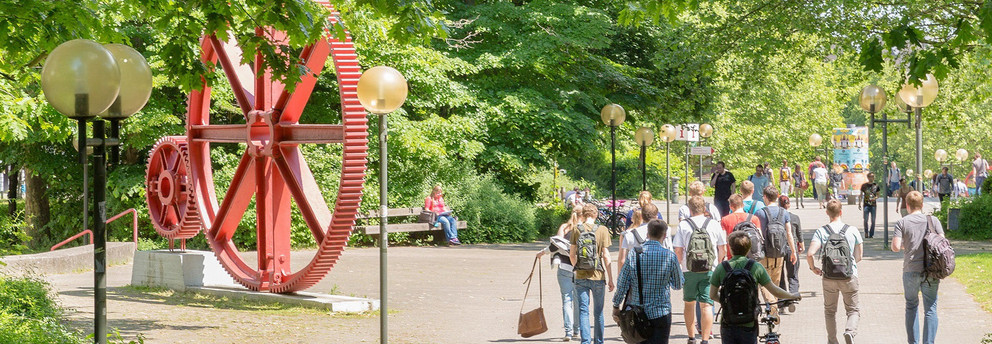 Studierende am Campus Nord Studierende laufen über den Campus Nord. Es ist eine große rote Skulptur in Form von großen Zahnrädern zu erkennen.
